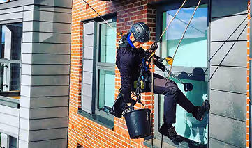 Worker wearing helmet and harness cleaning an exterior window while suspended on ropes from a brick and concrete building.