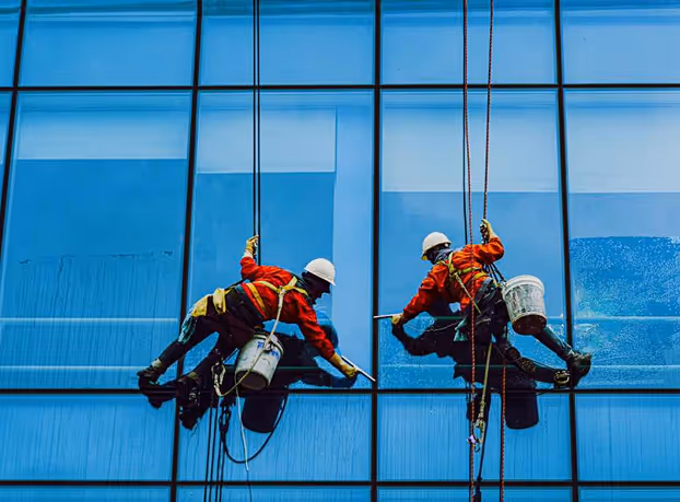 Two workers in safety gear cleaning glass windows on a high-rise building using ropes and buckets.