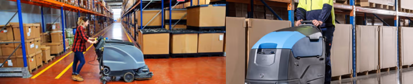 Two workers operating floor cleaning machines in a warehouse aisle lined with stacked cardboard boxes.