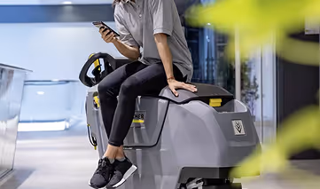 Person sitting on a grey floor-cleaning machine while looking at a smartphone.