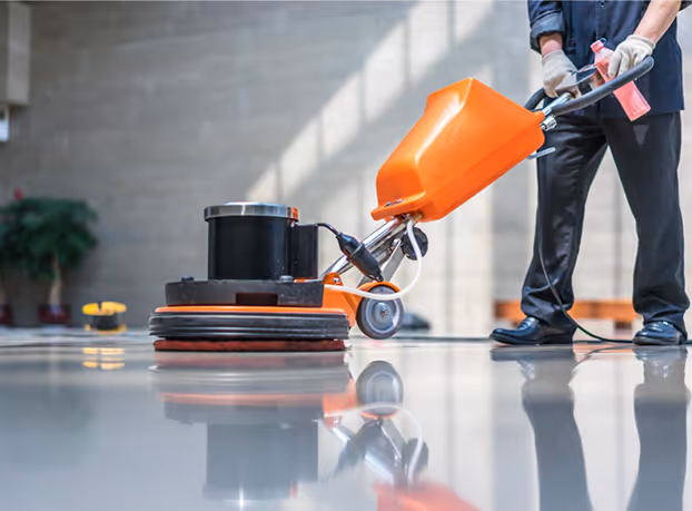 Person operating an orange floor polishing machine on a shiny indoor floor while holding a red spray bottle.