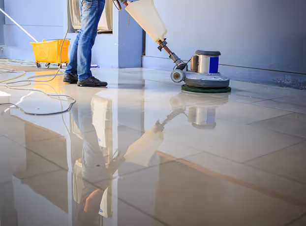 Person cleaning a shiny tile floor using a floor polishing machine with a yellow mop bucket nearby.