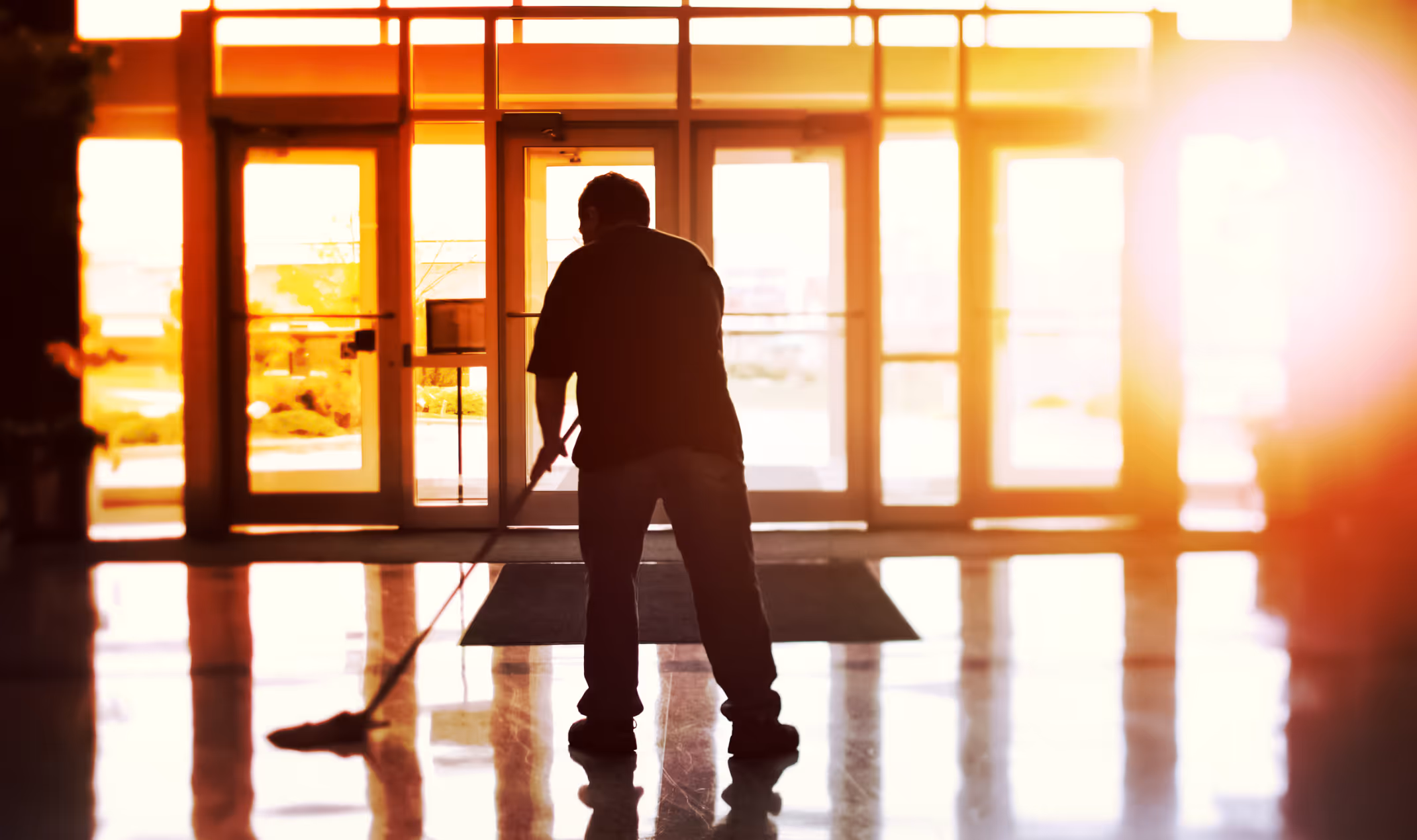 Silhouette of a person mopping the floor inside a building with large glass doors and bright sunlight outside.
