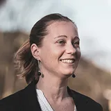 Portrait d'une femme souriante aux cheveux attachés en queue de cheval, avec un fond flou extérieur.