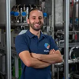 Homme souriant bras croisés dans une usine industrielle avec des tuyaux en acier en arrière-plan.
