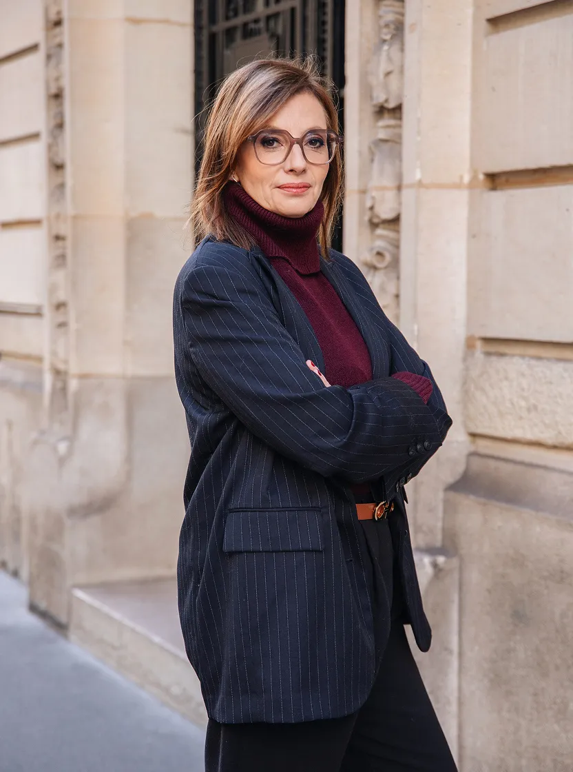 Femme aux cheveux bruns portant des lunettes, un pull à col roulé bordeaux et un blazer rayé bleu marine, posant avec les bras croisés devant un mur en pierre.