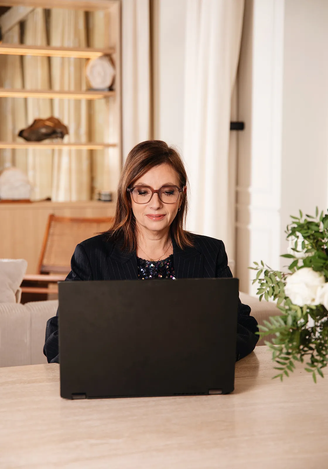 Femme portant des lunettes travaillant sur un ordinateur portable noir à une table avec des fleurs blanches à droite.