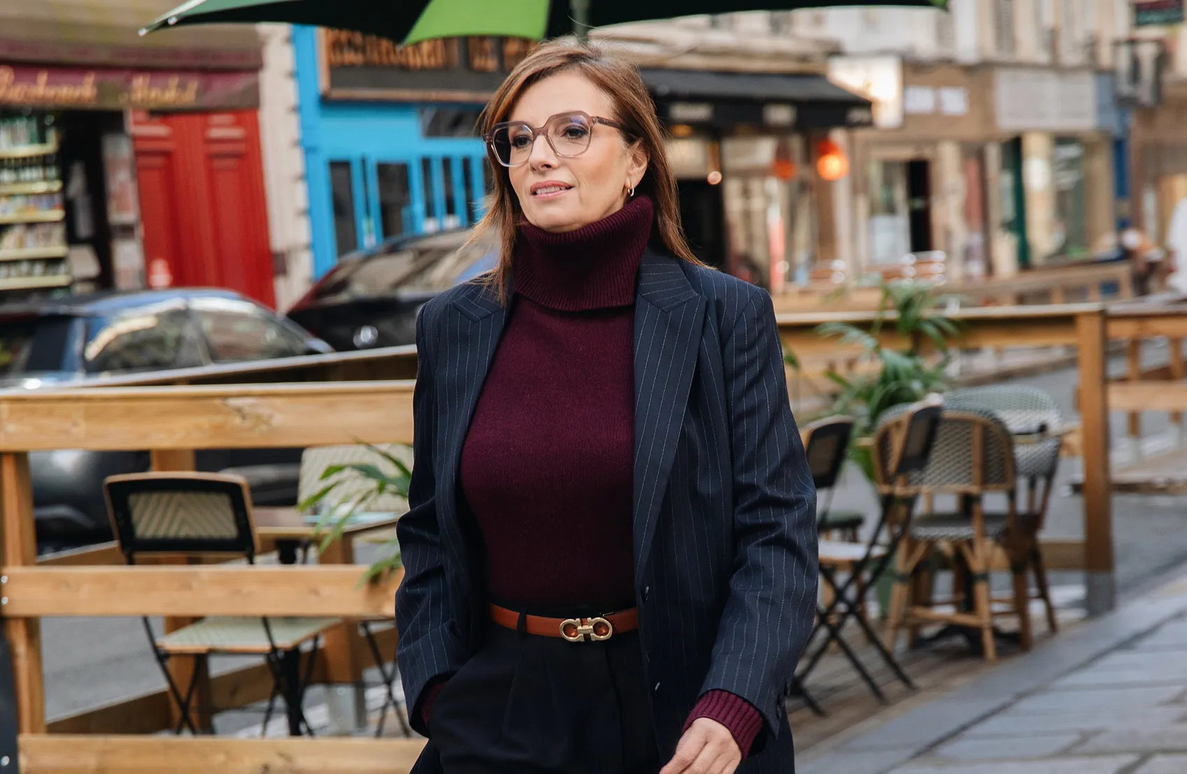 Femme portant un pull à col roulé bordeaux et une veste bleu marine à rayures marchant dans une rue avec des terrasses de café en arrière-plan.