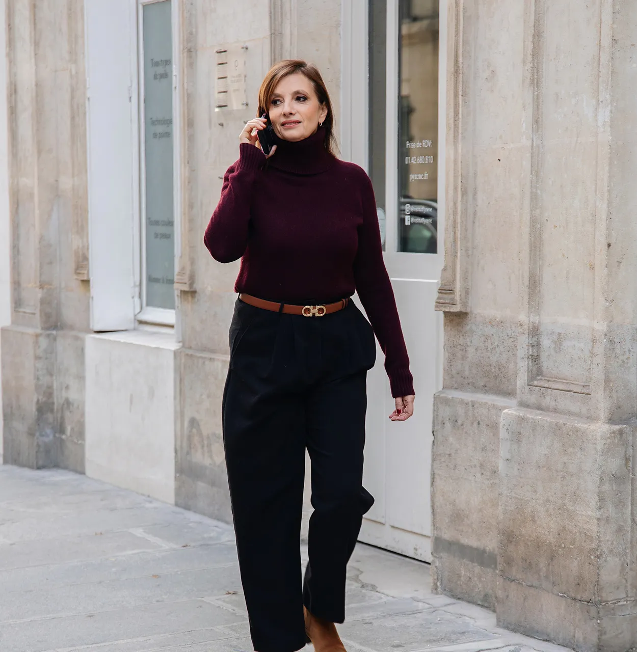 Femme marchant dans une rue urbaine tout en parlant au téléphone, portant un pull à col roulé bordeaux et un pantalon noir.