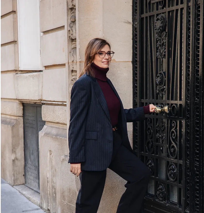 Femme souriante portant une veste à rayures et un col roulé, tenant la poignée d'une porte noire en fer forgé.