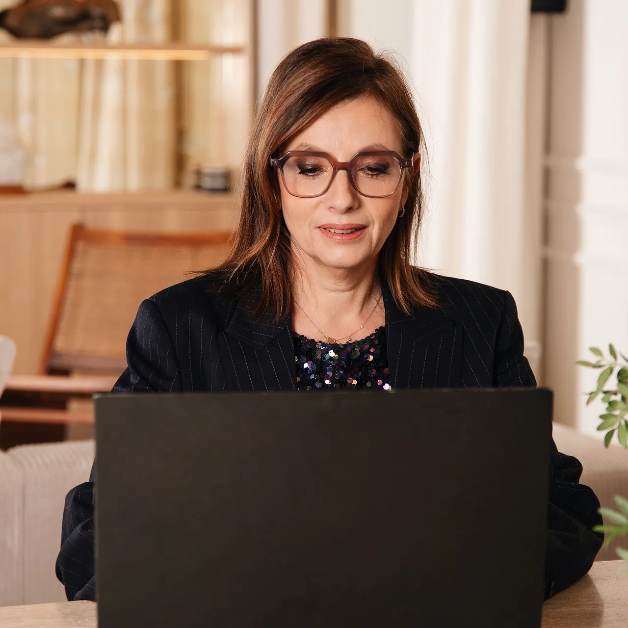 Femme portant des lunettes regardant attentivement un ordinateur portable noir dans un intérieur lumineux.