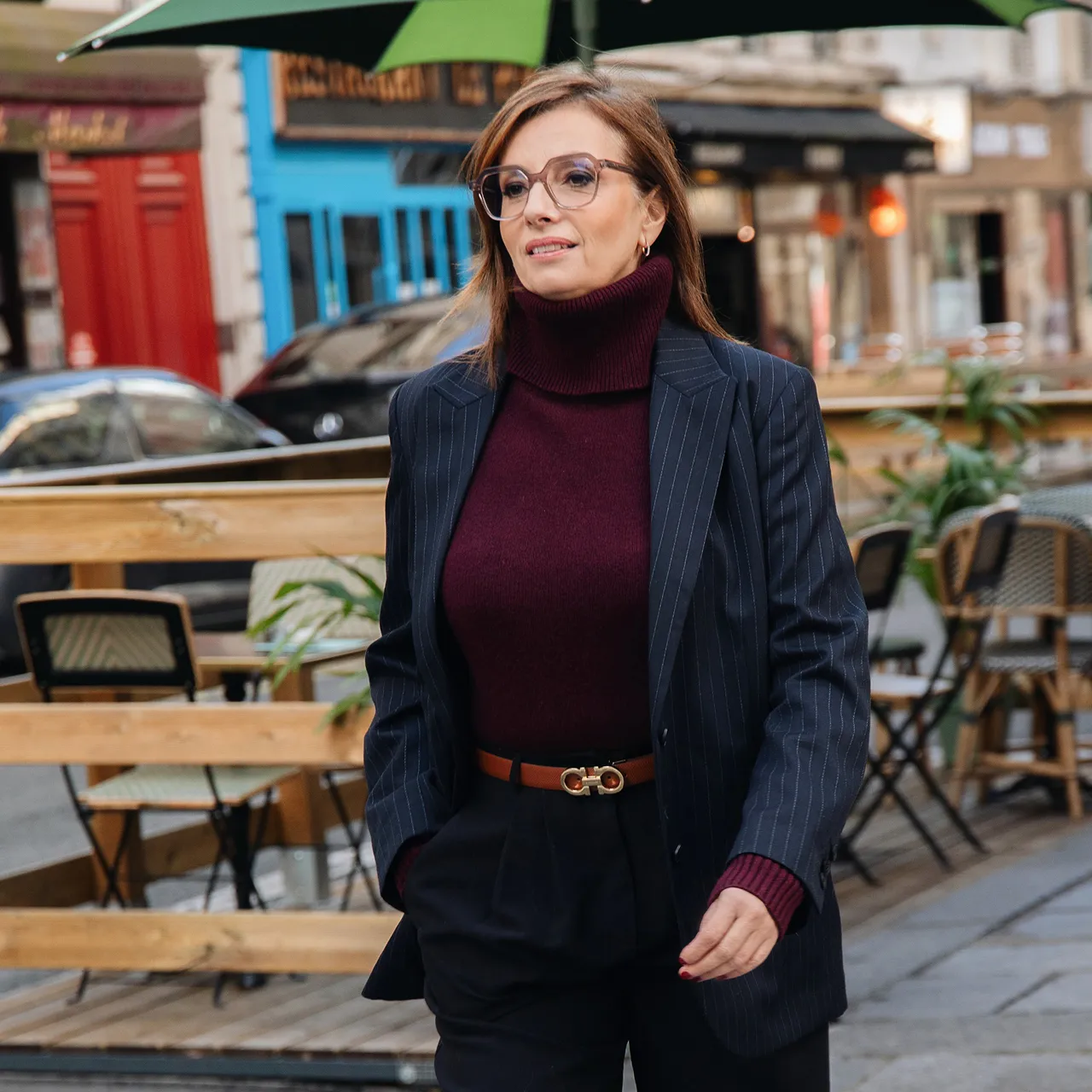 Femme portant un pull col roulé bordeaux et un blazer bleu marine à rayures marchant dans une rue avec un café en arrière-plan.