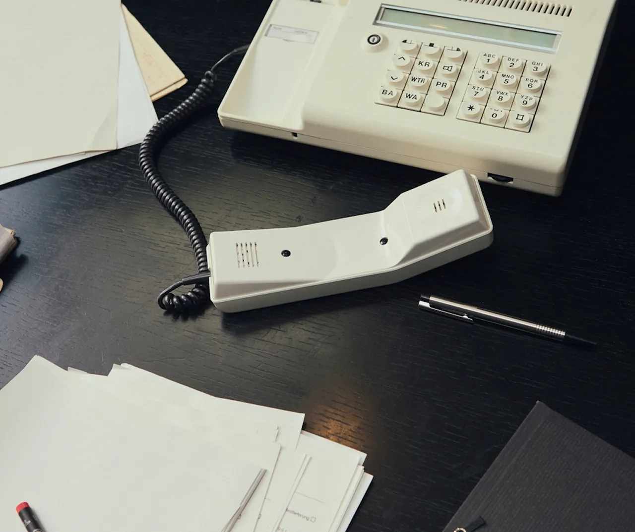 Téléphone de bureau blanc avec combiné posé sur une table noire entouré de papiers et d'un stylo.