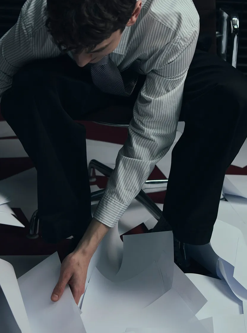Une personne en chemise à rayures et cravate ramasse des feuilles de papier éparpillées sur le sol.