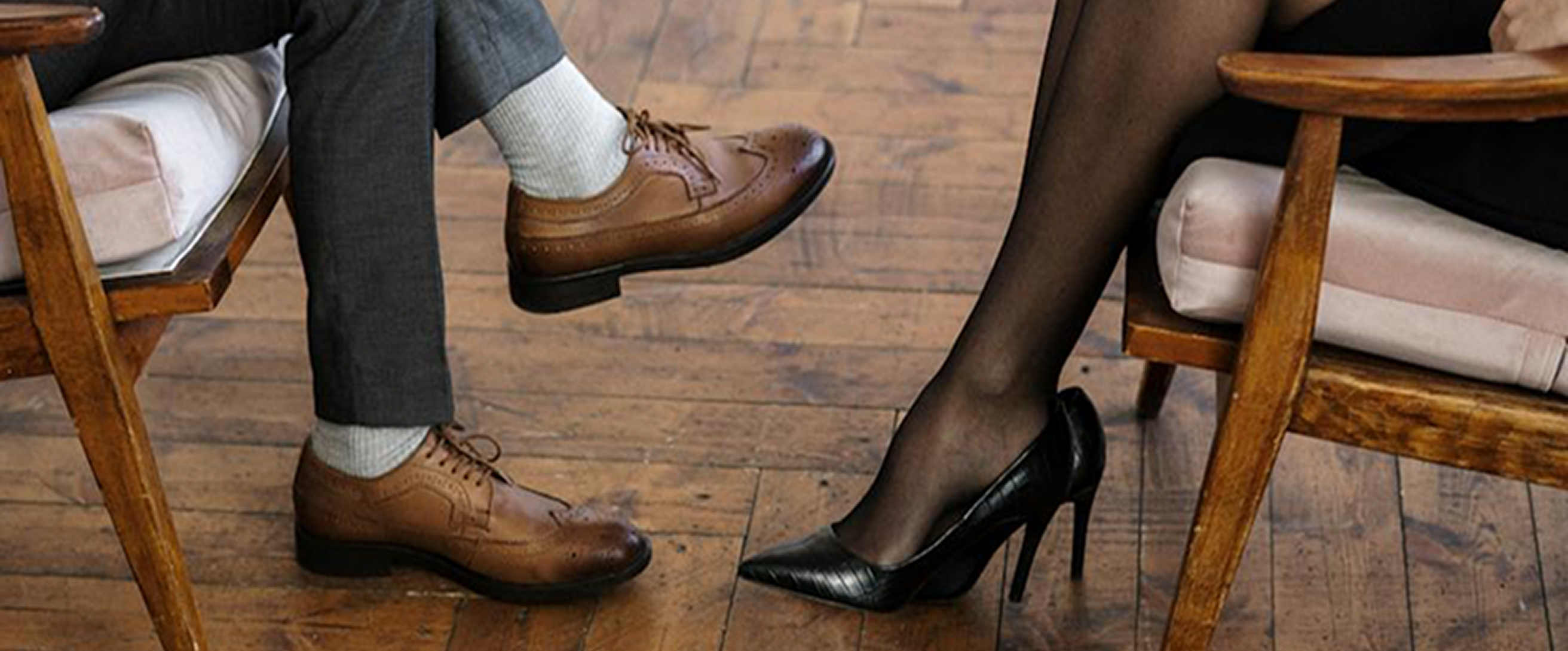 Pieds d'un homme en chaussures marron et pantalons gris croisées avec pieds d'une femme en collants noirs et talons noirs, assis sur des chaises en bois avec coussins.