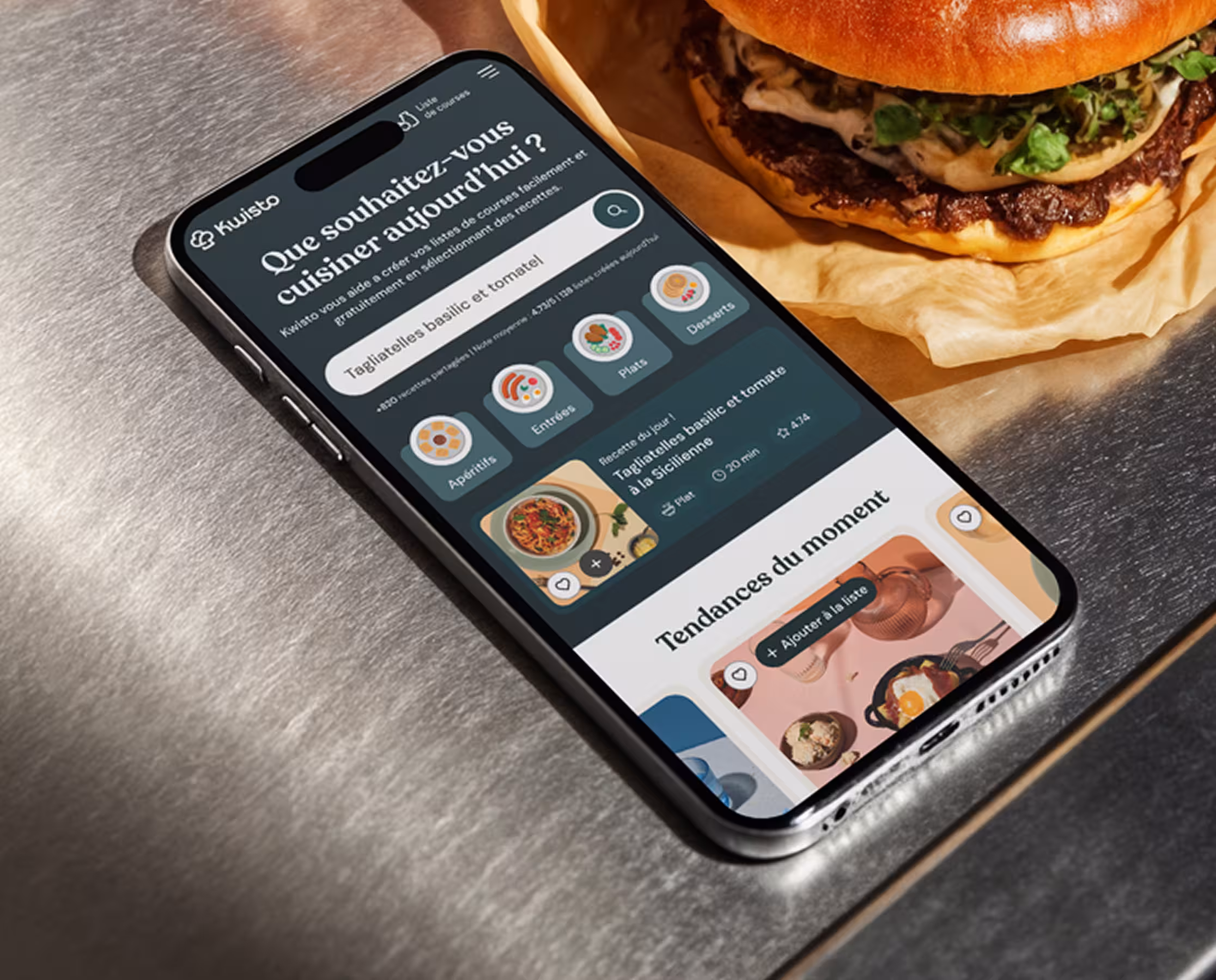 Smartphone affichant une interface d'application de cuisine française à côté d'un burger gourmet emballé sur une surface en métal.