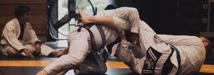 Two martial artists in white gi practicing a grappling technique on a mat with another person sitting in the background.