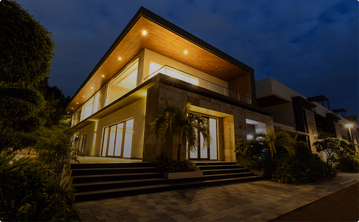 Modern two-story house at night with bright interior lights and plants by the entrance.