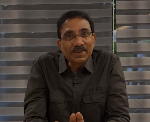 Man with glasses wearing a dark button-up shirt sitting in front of horizontal blinds.