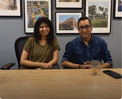Two people smiling while sitting at a conference table with framed photos on the wall behind them.