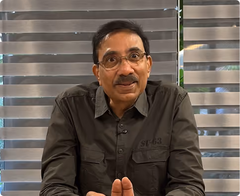 Man with glasses and mustache wearing a dark shirt sitting in front of window blinds.