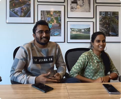 Two people sitting at a table with smartphones, framed photos on the wall behind them.