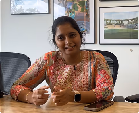 Smiling woman sitting at a table with a smartphone, framed artwork on the wall behind her.