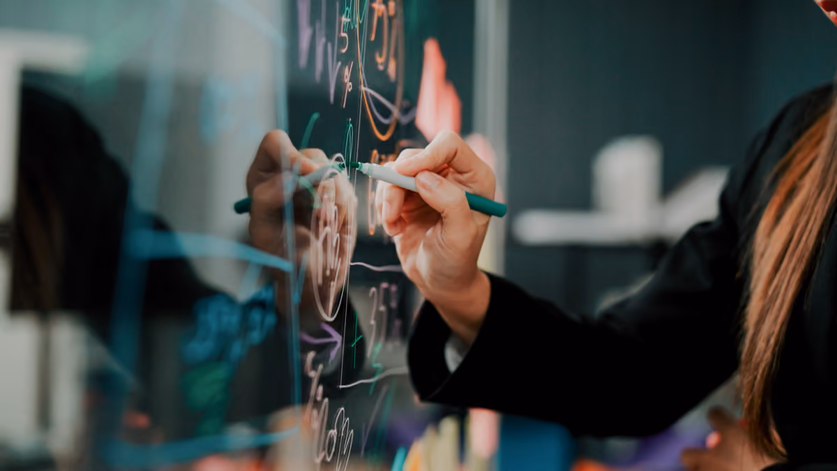 Person writing with a green marker on a glass board covered with colorful charts and numbers.