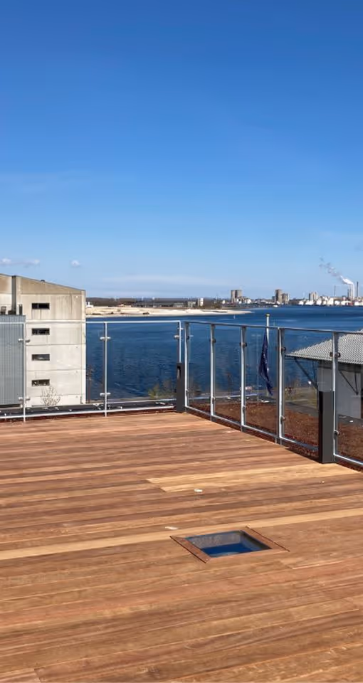Rummeligt terrassegulv i træ med glasværn og udsigt over havn, bygninger og klar blå himmel.