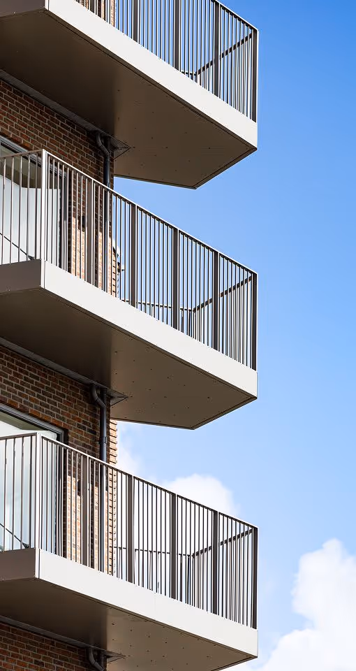 Tre hvide altaner med metalkonstruktion på en murstensbygning mod en blå himmel med få skyer.