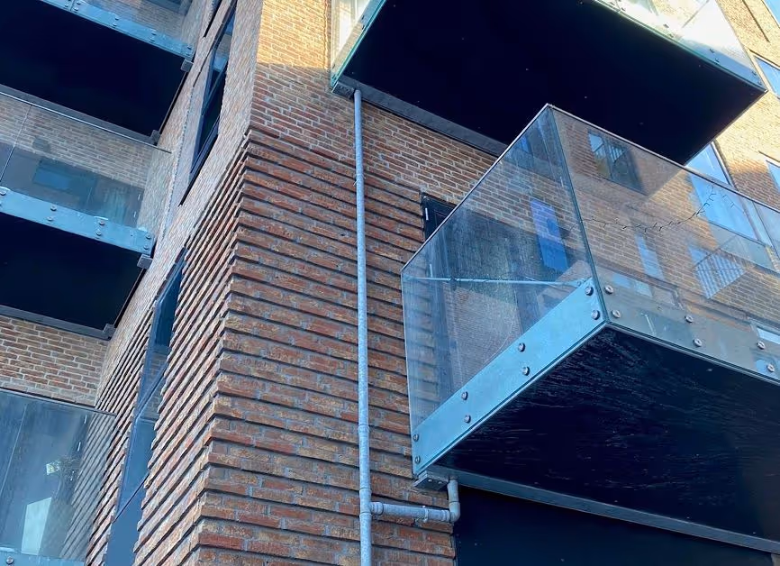 Nærbillede af et murstensbyggeri med glasaltaner og synlige metalrør langs hjørnet af bygningen.
