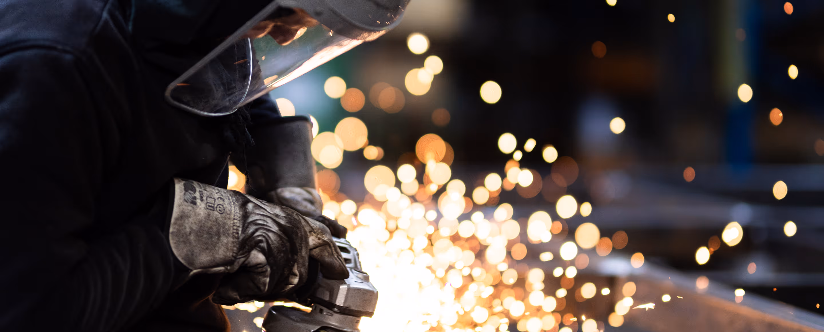 Person iført beskyttelsesudstyr sliber metal med vinkelsliber, gnister flyver omkring.