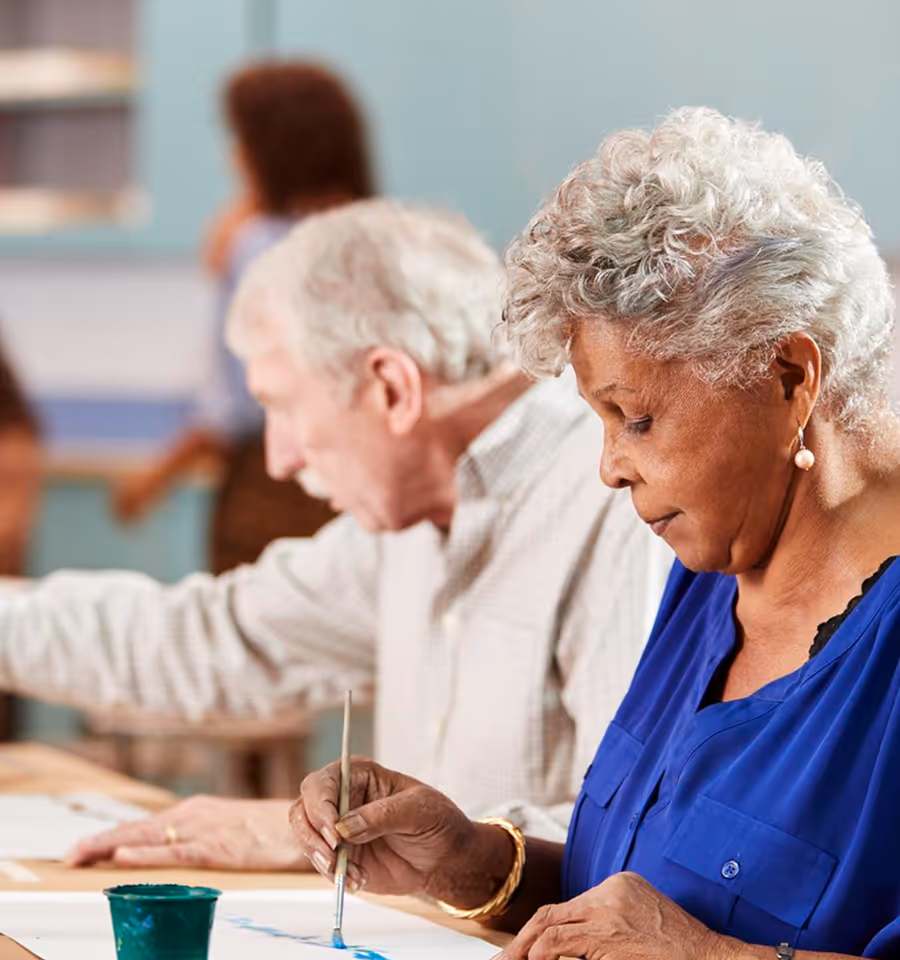 Elderly woman sitting and smiling while a caregiver in a purple shirt gently holds her arm and hand in a bright living space.