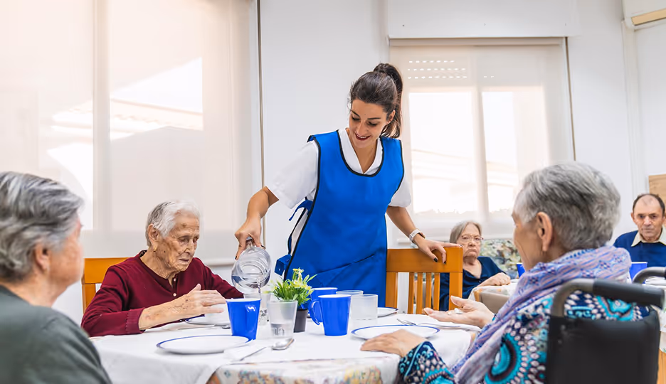 Caregiver wearing a blue apron pouring water for elderly people seated around a table in a bright room.