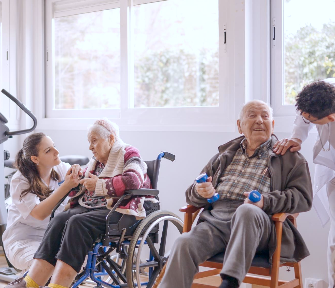 Caregivers interacting warmly with elderly man and woman in wheelchair during physical therapy session in a bright room.