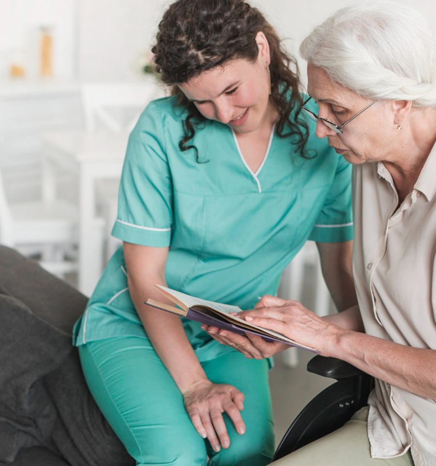 Young female caregiver in teal scrubs reading a book with an elderly woman in a wheelchair.