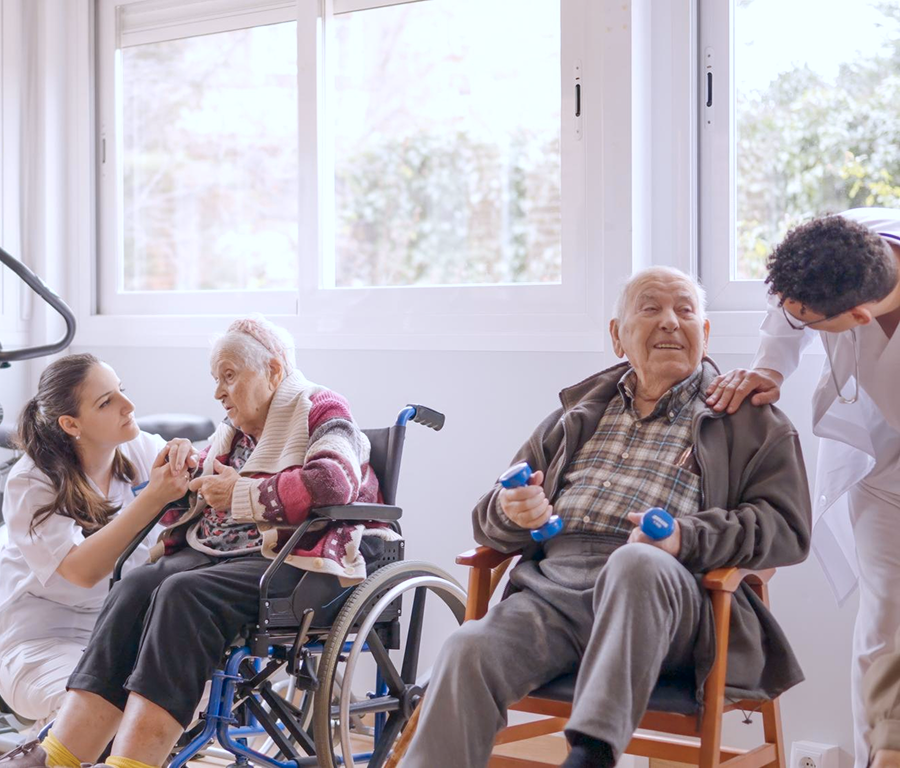 Two elderly people in a bright room with caregivers, one woman in a wheelchair holding hands with a nurse, and one man seated with light dumbbells, smiling at a standing caregiver.