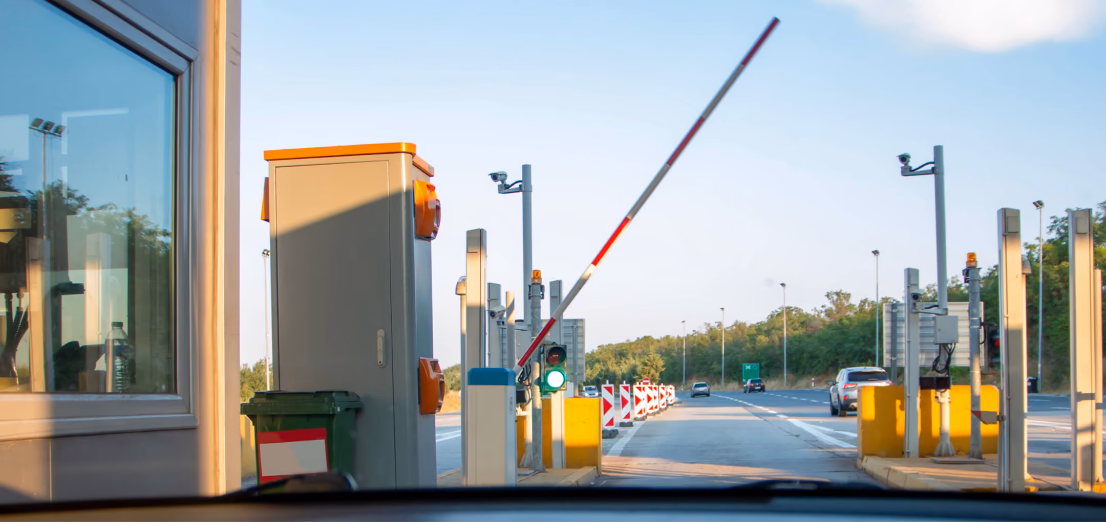 Raised barrier gate at a toll booth with green traffic light and multiple surveillance cameras on a highway lane.