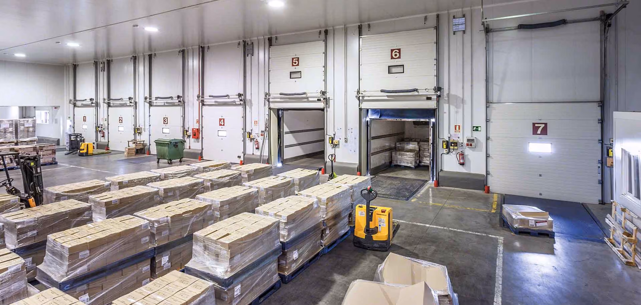 Warehouse loading dock area with pallets of boxed goods wrapped in plastic, forklifts, and dock doors numbered 1 through 7.