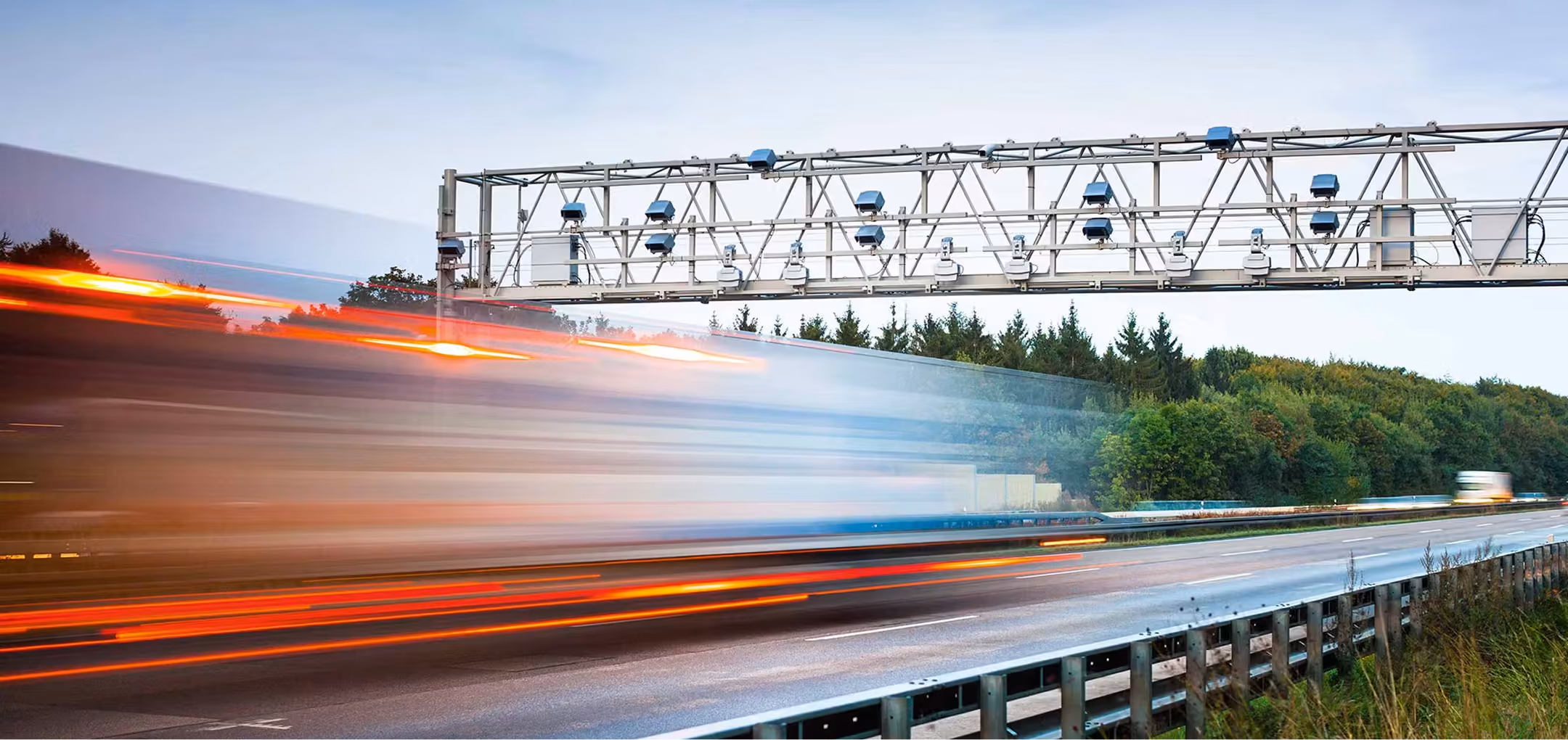 Blurred motion of a large truck speeding on a highway beneath an overhead gantry with multiple traffic monitoring cameras and sensors, bordered by green trees.
