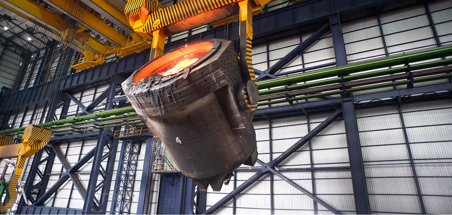 Large industrial ladle pouring molten metal inside a steel factory with metal beams and green pipes in the background.