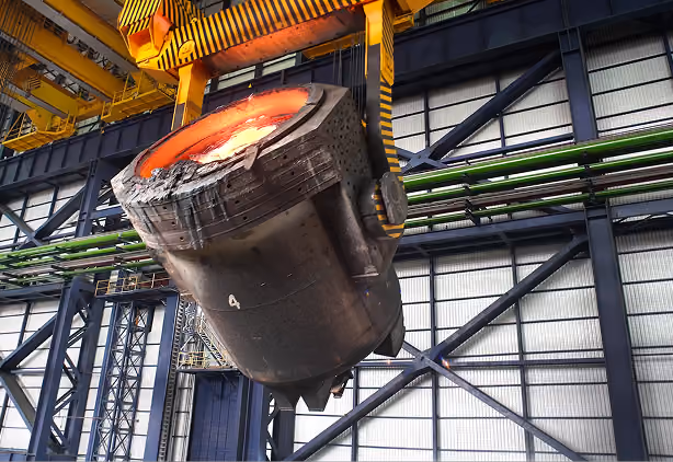 Large ladle vessel suspended by yellow overhead crane holding molten metal inside an industrial steel plant.
