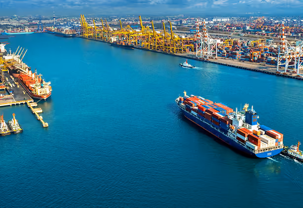 A large container ship loaded with colorful shipping containers navigating through a busy port with cranes and docked ships.