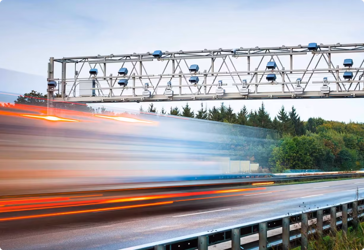 Blurred, fast-moving truck passing under a highway toll gantry with sensors, beside a wooded area.