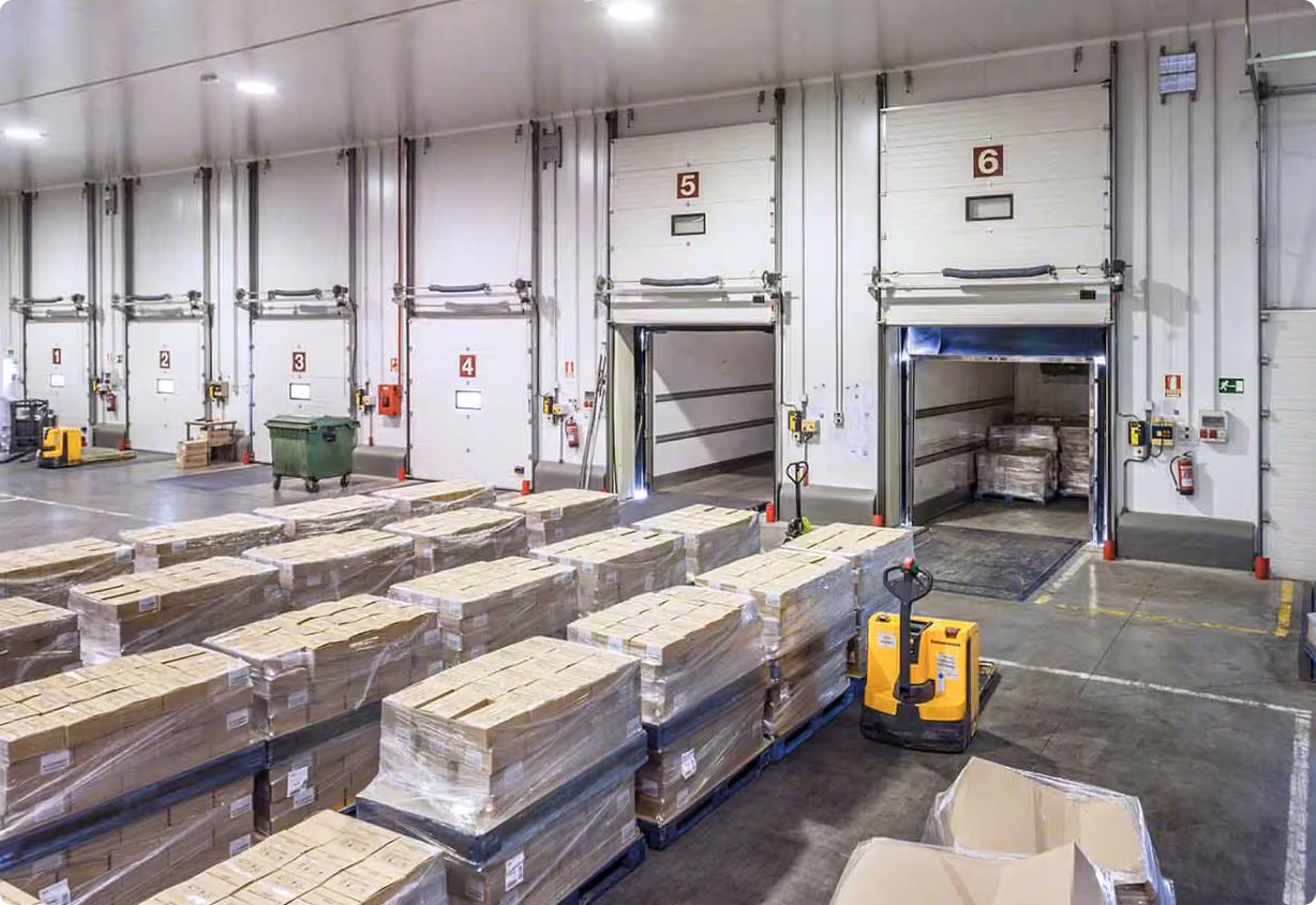Warehouse loading dock with multiple palletized boxes wrapped in plastic and two open dock doors labeled 5 and 6 for shipping and receiving.