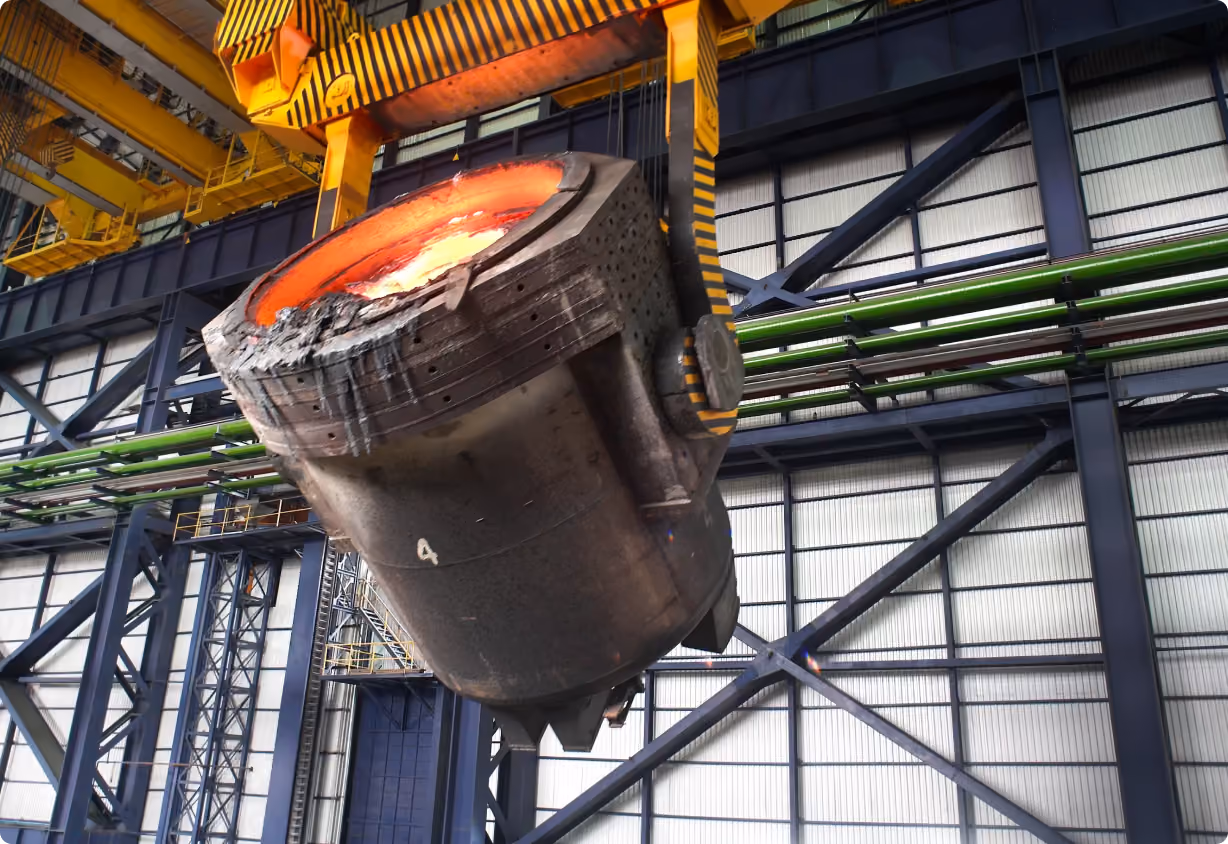 Large industrial ladle containing molten metal being lifted by a yellow overhead crane in a steel factory.