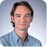 Portrait of a man with short brown hair wearing a light blue button-up shirt against a gray background.