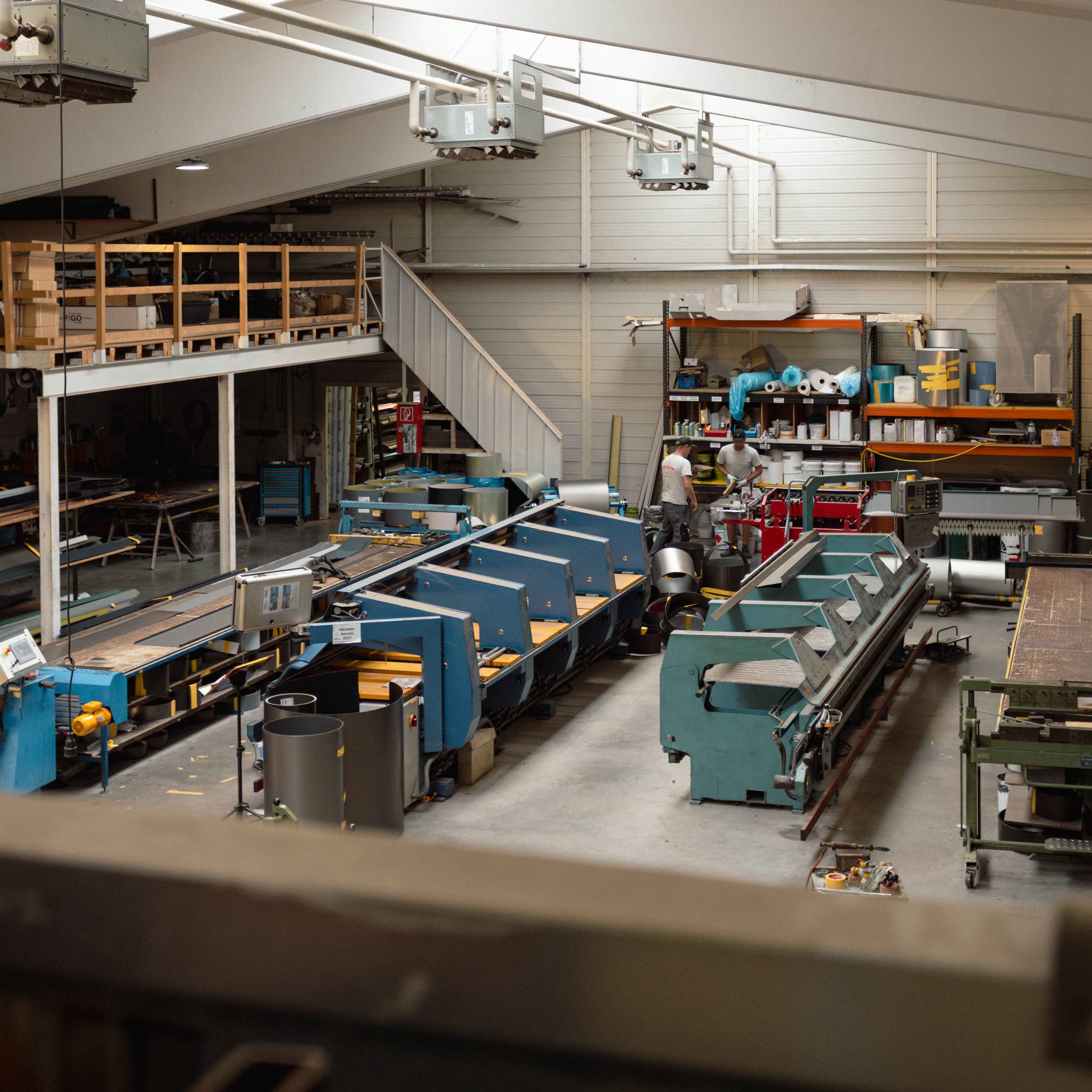 Industriehalle mit großen blauen Maschinen und zwei Arbeitern, die an einer Werkbank arbeiten.