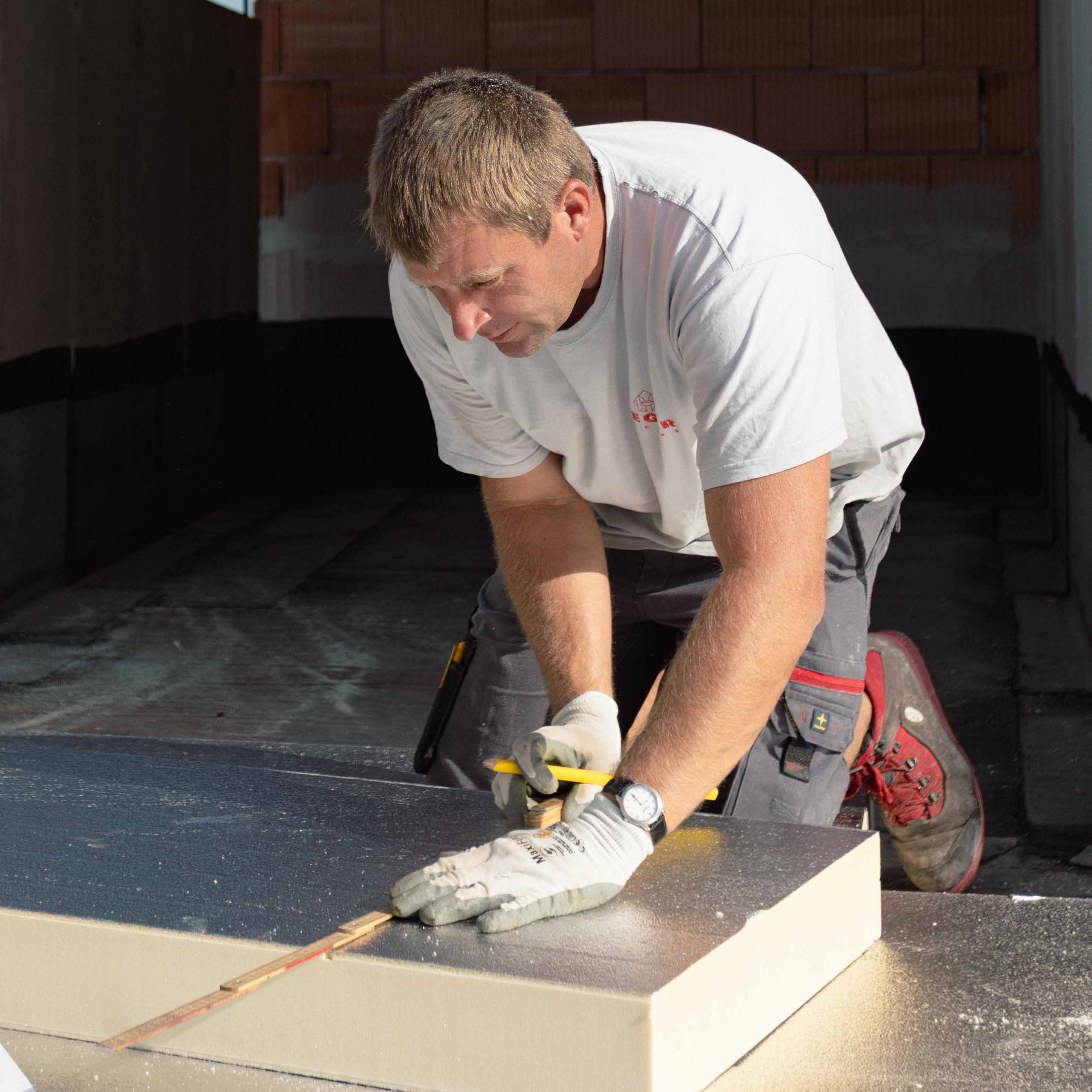 Bauarbeiter in weißem T-Shirt und grauer Hose kniet und markiert eine Dämmplatte mit einem Bleistift.