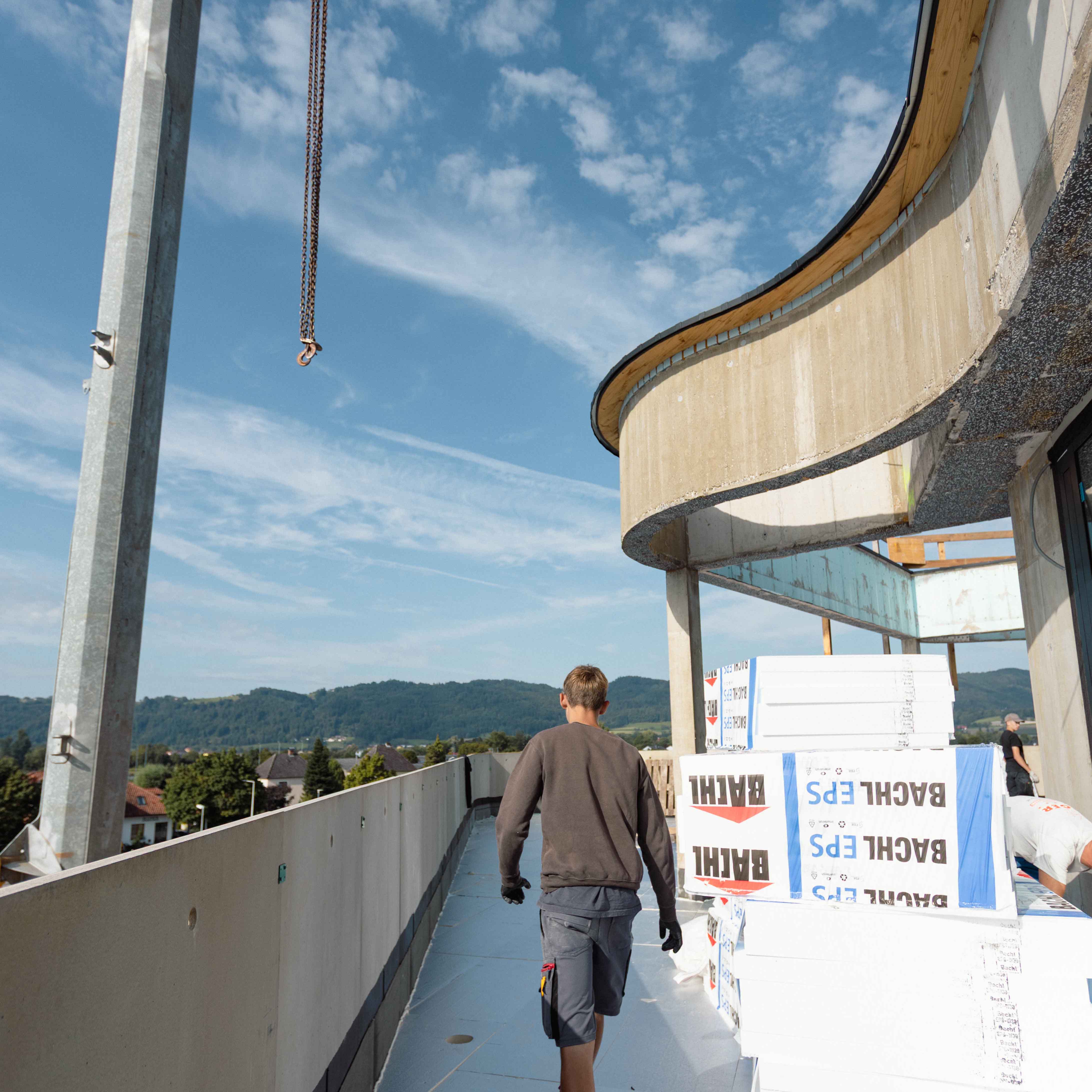 Bauarbeiter mit dunkelgrauem Pullover und Shorts läuft auf einer Baustelle auf einem Balkon mit Bauholz und einer gebogenen Betonbalustrade unter blauem Himmel.